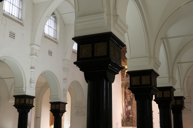 The image depicts the interior of a historic building with ornate, dark wooden columns topped with decorative capitals. The walls are painted white, and there are high, arched windows allowing soft natural light to illuminate the space. The architecture features a series of arches and detailed moldings.