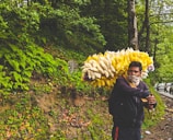 A vendor stands by the roadside in a lush, green forest area, carrying a big bundle of packets filled with snacks. The man is wearing a jacket and has his face partially covered with a cloth. Several cars are lined up along the road, surrounded by dense trees and ferns.