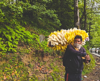 A vendor stands by the roadside in a lush, green forest area, carrying a big bundle of packets filled with snacks. The man is wearing a jacket and has his face partially covered with a cloth. Several cars are lined up along the road, surrounded by dense trees and ferns.
