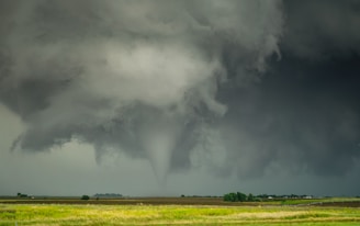 A large tornado funnel cloud descends from stormy, dark clouds over a flat, rural landscape with fields and scattered buildings.