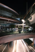 A nighttime urban scene featuring elevated roadways and walkways with light trails from passing vehicles below. The infrastructure includes modern design elements like curved bridges and a glass railing on the pedestrian level. Potted plants add greenery to the scene, and streetlights create a well-lit environment.