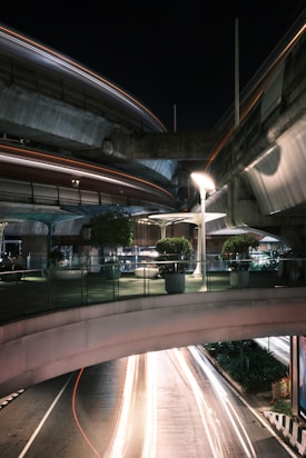 A nighttime urban scene featuring elevated roadways and walkways with light trails from passing vehicles below. The infrastructure includes modern design elements like curved bridges and a glass railing on the pedestrian level. Potted plants add greenery to the scene, and streetlights create a well-lit environment.