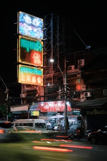 A vibrant street scene at night with neon signs featuring Asian characters, and a variety of colors including blue, red, and yellow. Below the signs is a building with multiple businesses, including a food stall with a brightly lit red awning. Cars and motorcycles create motion blur as they pass by on the street lined with power cables.