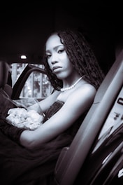 A person with long braided hair is seated elegantly inside a car, holding a bouquet of white flowers. They are wearing a pearl necklace and a dark strapless dress with gloved arms, creating a classic and sophisticated appearance.