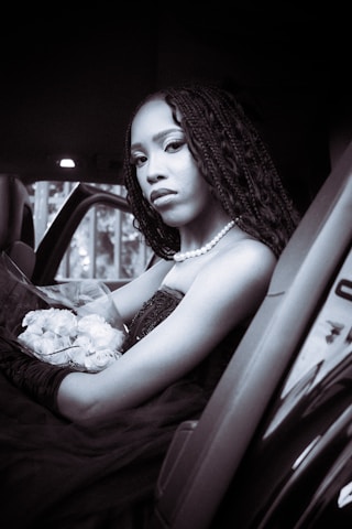 A person with long braided hair is seated elegantly inside a car, holding a bouquet of white flowers. They are wearing a pearl necklace and a dark strapless dress with gloved arms, creating a classic and sophisticated appearance.