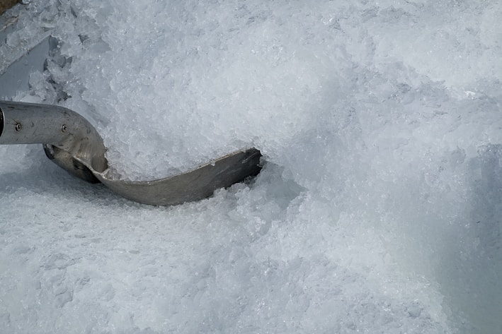 A large pile of crushed ice with a metal scoop partially buried in it. The ice appears translucent and frosty, with varying shades and textures giving a sense of coldness.