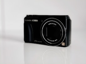 A digital camera with a black body, displaying model details and lens specifications on the front. The lens is prominent, surrounded by branding text, and the camera sits on a reflective white surface against a plain background.