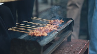 A rustic grill holds several skewers of marinated meat, cooking over open flames. Smoke rises gently from the meat, indicating it's being grilled. The scene seems outdoors, with people possibly gathered around.