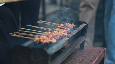 A rustic grill holds several skewers of marinated meat, cooking over open flames. Smoke rises gently from the meat, indicating it's being grilled. The scene seems outdoors, with people possibly gathered around.