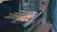 A rustic grill holds several skewers of marinated meat, cooking over open flames. Smoke rises gently from the meat, indicating it's being grilled. The scene seems outdoors, with people possibly gathered around.