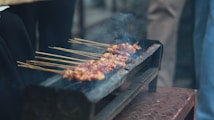 A rustic grill holds several skewers of marinated meat, cooking over open flames. Smoke rises gently from the meat, indicating it's being grilled. The scene seems outdoors, with people possibly gathered around.