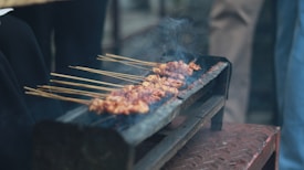 A rustic grill holds several skewers of marinated meat, cooking over open flames. Smoke rises gently from the meat, indicating it's being grilled. The scene seems outdoors, with people possibly gathered around.