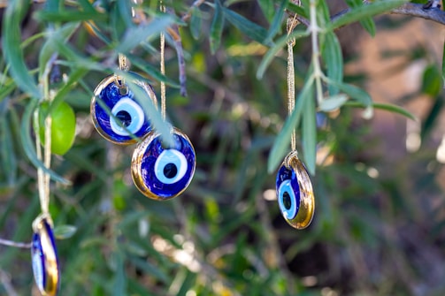 Hanging on the branches of a tree are several blue and white glass charms with eye motifs, traditionally known as 'evil eye' talismans. The surrounding foliage is green and lush, creating a natural and serene backdrop.