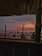 A beach scene viewed from a shaded structure, featuring people gathered along the shoreline. The sky exhibits a dramatic sunset with vivid hues of orange, pink, and purple. The foreground includes a roof made of bamboo or wooden slats, with silhouettes of individuals sitting and standing as they gaze towards the ocean.