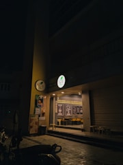 An exterior view of a tea shop at night with a dimly lit entrance. The storefront features a circular lit sign with the shop’s logo, promotional posters on the wall, and glass windows through which interior seating and decor with framed quotes can be seen. There are stools placed outside and several scooters parked nearby.