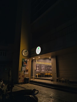An exterior view of a tea shop at night with a dimly lit entrance. The storefront features a circular lit sign with the shop’s logo, promotional posters on the wall, and glass windows through which interior seating and decor with framed quotes can be seen. There are stools placed outside and several scooters parked nearby.