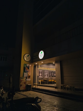 An exterior view of a tea shop at night with a dimly lit entrance. The storefront features a circular lit sign with the shop&rsquo;s logo, promotional posters on the wall, and glass windows through which interior seating and decor with framed quotes can be seen. There are stools placed outside and several scooters parked nearby.