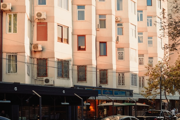 A residential building with beige-colored walls and multiple windows, some of which have air conditioning units attached. The lower part of the building features shops or cafes with awnings. Cars and people are visible on the street, and there are trees on the right side.