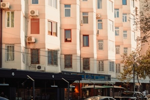 A residential building with beige-colored walls and multiple windows, some of which have air conditioning units attached. The lower part of the building features shops or cafes with awnings. Cars and people are visible on the street, and there are trees on the right side.