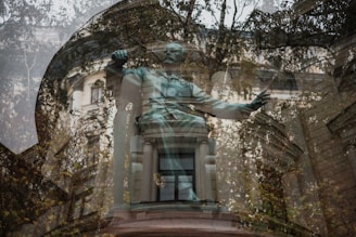 A reflective composition showing a statue overlaid with the image of a building facade and trees. The statue is captured with an arm raised, and the building in the background features classical architectural elements such as columns. The trees add a natural element, blending with the structure and creating an ethereal effect.
