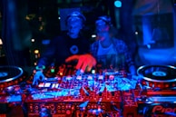 Two DJs wearing headphones are operating a mixing console with vibrant lighting. The scene includes various buttons and knobs on the console, with a multitude of colorful lights reflecting off the equipment.