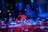 Two DJs wearing headphones are operating a mixing console with vibrant lighting. The scene includes various buttons and knobs on the console, with a multitude of colorful lights reflecting off the equipment.