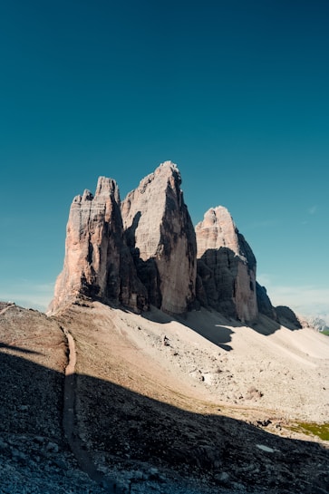 A breathtaking view of towering mountain peaks with rugged, rocky surfaces standing against a deep blue sky. A narrow path leads towards the base of the dramatic rock formations, and the ground is covered in loose stones and sparse vegetation.