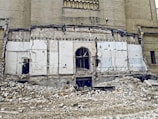A partially demolished building with exposed brickwork and remnants of a wall. Rubble and debris cover the ground, and there are visible cords and structural elements. The scene suggests ongoing demolition or abandonment.