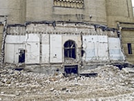 A partially demolished building with exposed brickwork and remnants of a wall. Rubble and debris cover the ground, and there are visible cords and structural elements. The scene suggests ongoing demolition or abandonment.