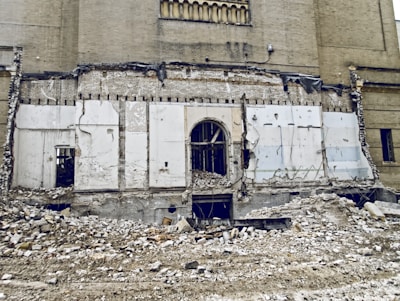 A partially demolished building with exposed brickwork and remnants of a wall. Rubble and debris cover the ground, and there are visible cords and structural elements. The scene suggests ongoing demolition or abandonment.