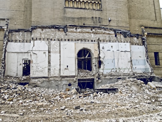 A partially demolished building with exposed brickwork and remnants of a wall. Rubble and debris cover the ground, and there are visible cords and structural elements. The scene suggests ongoing demolition or abandonment.