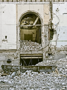 A heavily damaged building wall with a large arched opening surrounded by debris. The structure appears to be partially collapsed, exposing internal elements like pipes and wires. Various materials, including bricks and metal, are scattered across the ground.