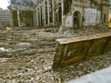 A partially demolished building surrounded by rubble and debris. The structure has missing walls and exposed beams, with piles of bricks and dirt scattered around. A large metal blade, possibly from a bulldozer, is positioned in front of the scene.