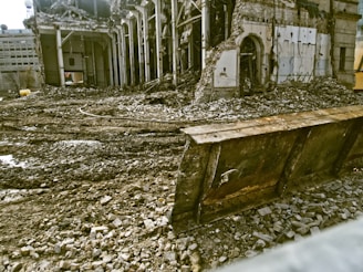 A partially demolished building surrounded by rubble and debris. The structure has missing walls and exposed beams, with piles of bricks and dirt scattered around. A large metal blade, possibly from a bulldozer, is positioned in front of the scene.