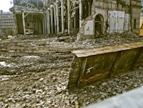 A partially demolished building surrounded by rubble and debris. The structure has missing walls and exposed beams, with piles of bricks and dirt scattered around. A large metal blade, possibly from a bulldozer, is positioned in front of the scene.