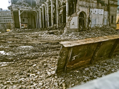 A partially demolished building surrounded by rubble and debris. The structure has missing walls and exposed beams, with piles of bricks and dirt scattered around. A large metal blade, possibly from a bulldozer, is positioned in front of the scene.
