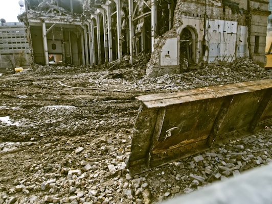 A partially demolished building surrounded by rubble and debris. The structure has missing walls and exposed beams, with piles of bricks and dirt scattered around. A large metal blade, possibly from a bulldozer, is positioned in front of the scene.