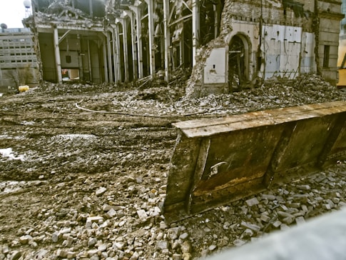 A partially demolished building surrounded by rubble and debris. The structure has missing walls and exposed beams, with piles of bricks and dirt scattered around. A large metal blade, possibly from a bulldozer, is positioned in front of the scene.