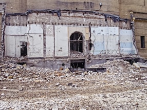 A partially demolished brick building reveals its interior structure, with debris and rubble scattered across the foreground. The remnants of walls with exposed beams and wires are visible, along with a large doorway at the center.