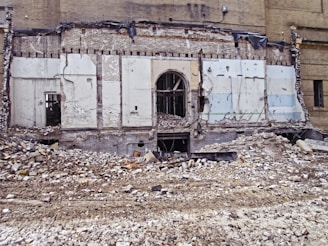A partially demolished brick building reveals its interior structure, with debris and rubble scattered across the foreground. The remnants of walls with exposed beams and wires are visible, along with a large doorway at the center.