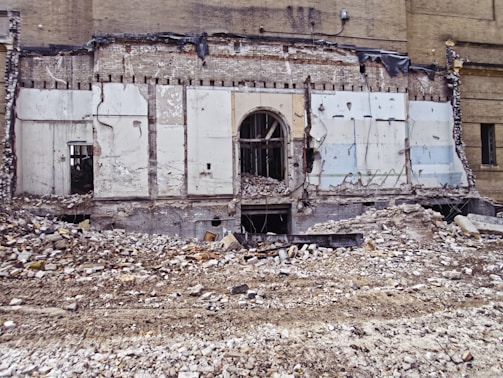 A partially demolished brick building reveals its interior structure, with debris and rubble scattered across the foreground. The remnants of walls with exposed beams and wires are visible, along with a large doorway at the center.