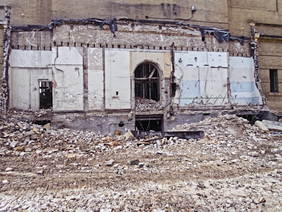 A partially demolished brick building reveals its interior structure, with debris and rubble scattered across the foreground. The remnants of walls with exposed beams and wires are visible, along with a large doorway at the center.
