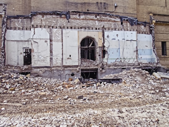 A partially demolished brick building reveals its interior structure, with debris and rubble scattered across the foreground. The remnants of walls with exposed beams and wires are visible, along with a large doorway at the center.