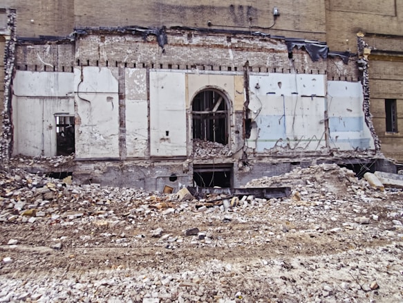 A partially demolished brick building reveals its interior structure, with debris and rubble scattered across the foreground. The remnants of walls with exposed beams and wires are visible, along with a large doorway at the center.