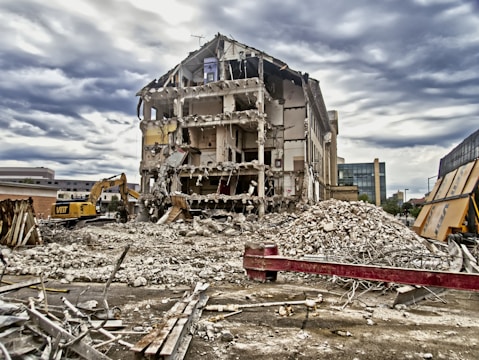 A heavily damaged building with several floors partially or completely destroyed. Debris is scattered across the ground, and a large construction vehicle is present, likely involved in demolition work. The sky is overcast, creating a dramatic background.