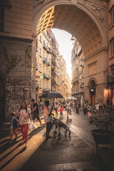 A bustling urban street scene with people walking and engaging in activities. The street is framed by an architectural archway, casting shadows on the cobblestone pavement. Sunlight filters through, creating a warm and inviting atmosphere. Buildings line both sides of the street, with some balconies adorned with plants. Shoppers, families, and individuals with strollers populate the area, suggesting a lively and vibrant city life.