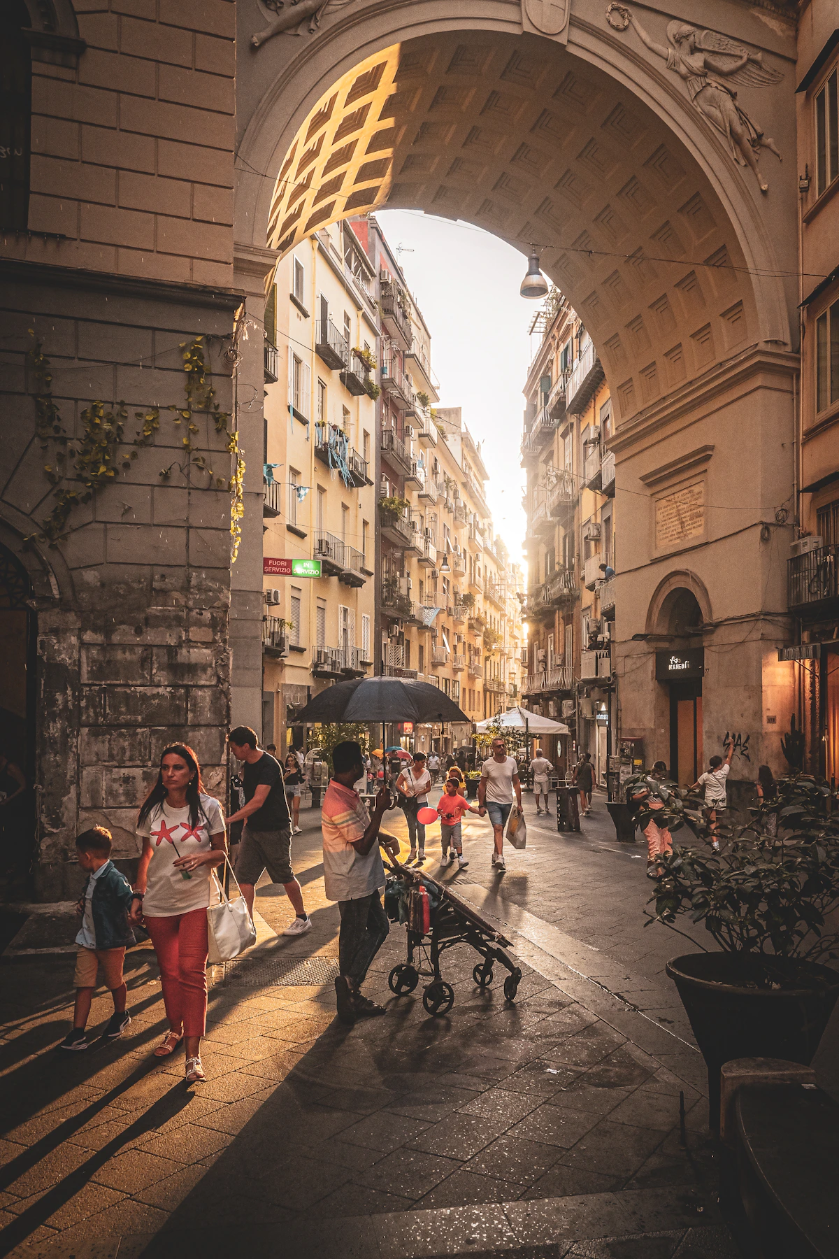 Families and pedestrians beneath an arch on a lively Naples street in warm evening light