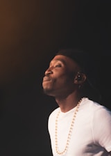 A person wearing a white shirt, a black cap, and a beaded necklace is looking upwards with a contemplative expression. The background is dark and the lighting highlights the person's face.