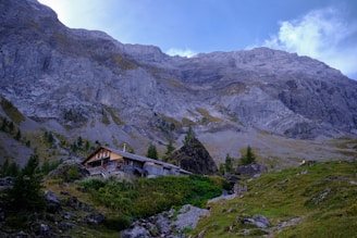 A rustic wooden cabin is nestled among greenery and rugged terrain at the base of towering gray limestone mountains. Sparse trees and shrubs dot the gentle slopes leading up to the majestic cliffs, creating a serene and isolated setting. The sky above is partly cloudy with patches of bright blue.