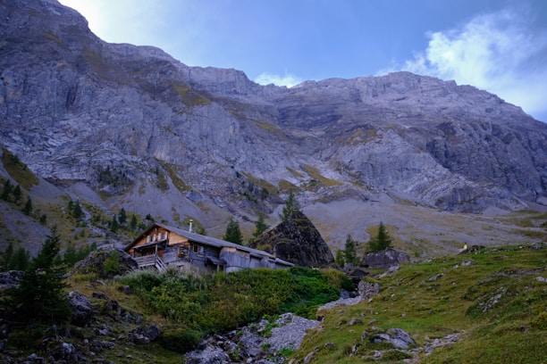 A rustic wooden cabin is nestled among greenery and rugged terrain at the base of towering gray limestone mountains. Sparse trees and shrubs dot the gentle slopes leading up to the majestic cliffs, creating a serene and isolated setting. The sky above is partly cloudy with patches of bright blue.